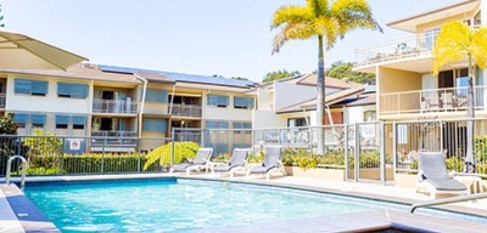 A resort-style swimming pool and hot tub are surrounded by lounge chairs and palm trees, with apartment buildings in the background under a clear blue sky.