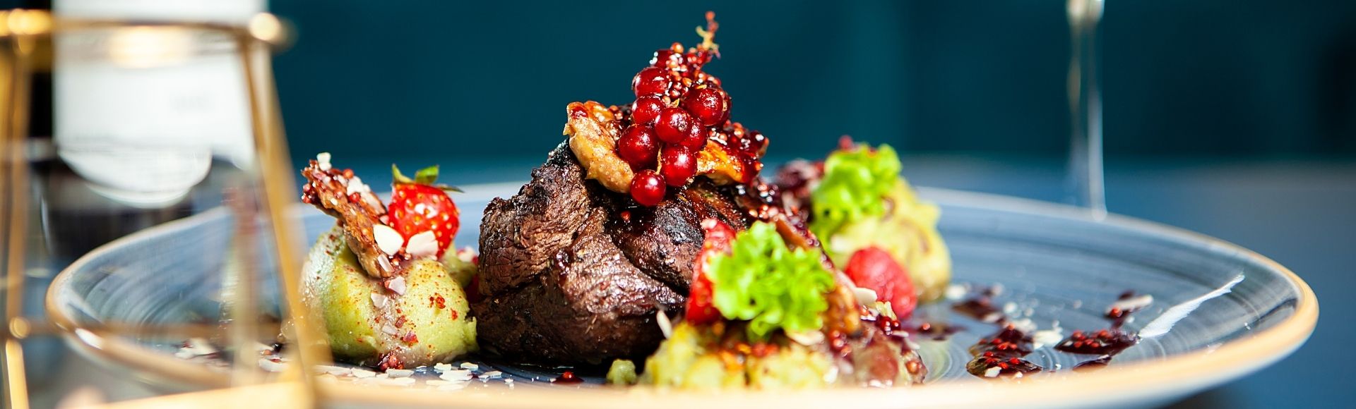 A gourmet dish featuring a seared steak topped with red currants, garnished with decorative greens and vibrant sauces, set on an elegant plate.