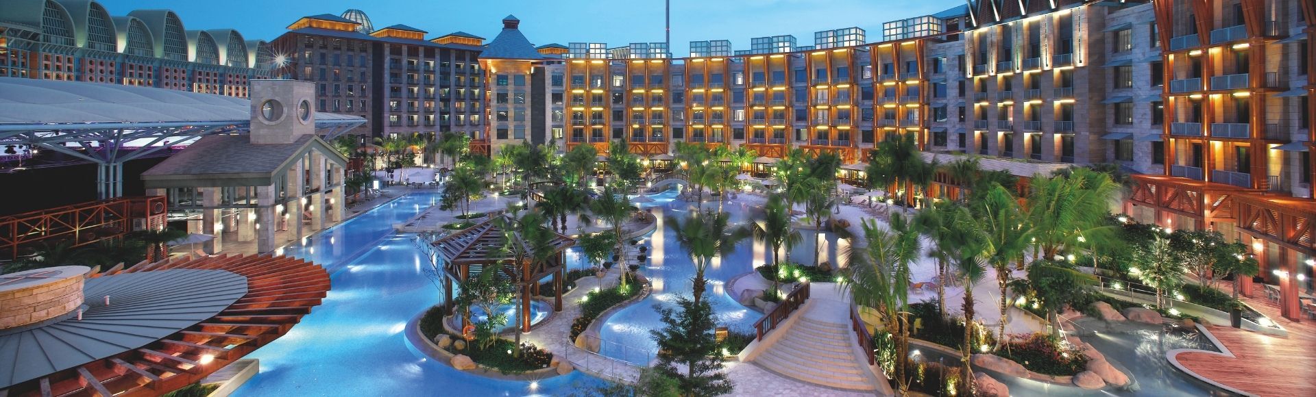 Luxurious resort scene at dusk with a winding pool, lush greenery, palm trees, and warmly lit modern buildings, evoking a sense of tropical elegance.