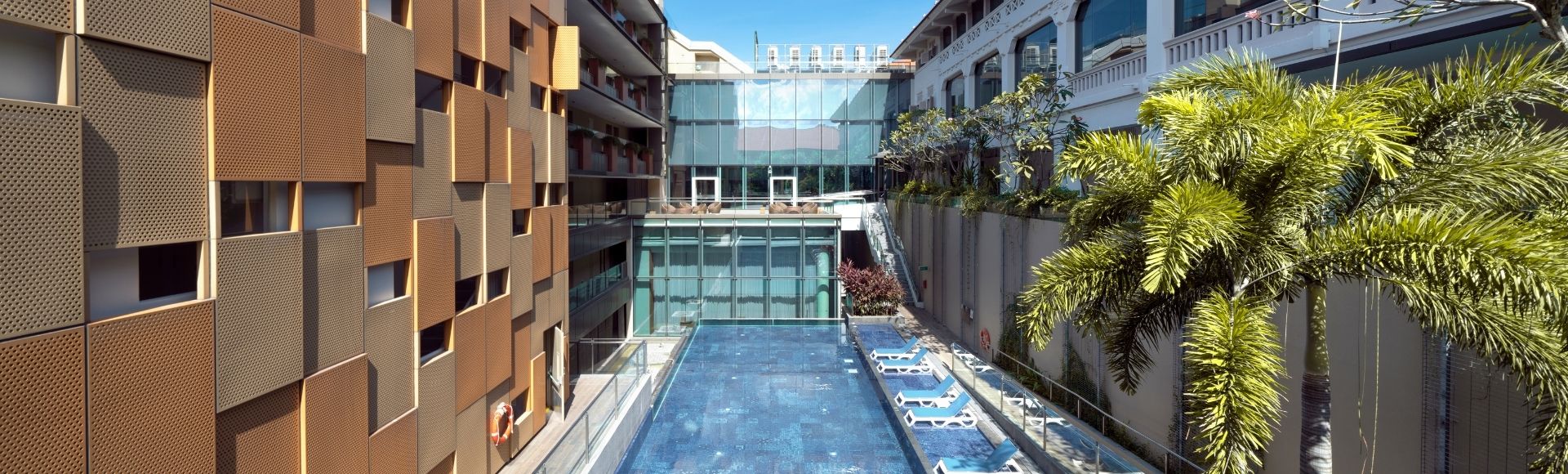Rooftop view of a modern hotel featuring a central swimming pool, flanked by tall buildings with honeycomb-patterned walls and lush palm trees.