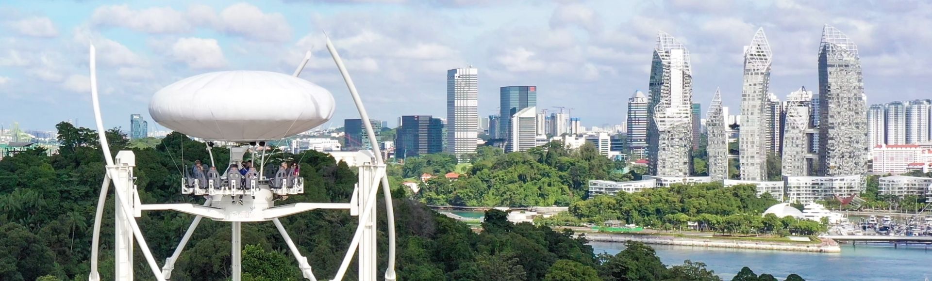 Futuristic pod ride elevated above lush green trees with cityscape in the background, including unique, reflective skyscrapers under a blue sky.