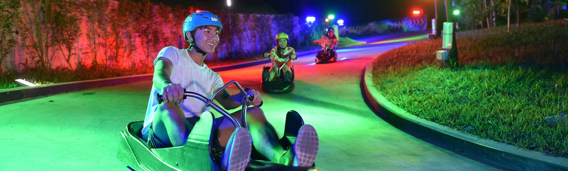 A person enjoys riding a luge cart down a winding track at night, illuminated by colorful lights. They wear a helmet and smile, capturing a sense of fun.
