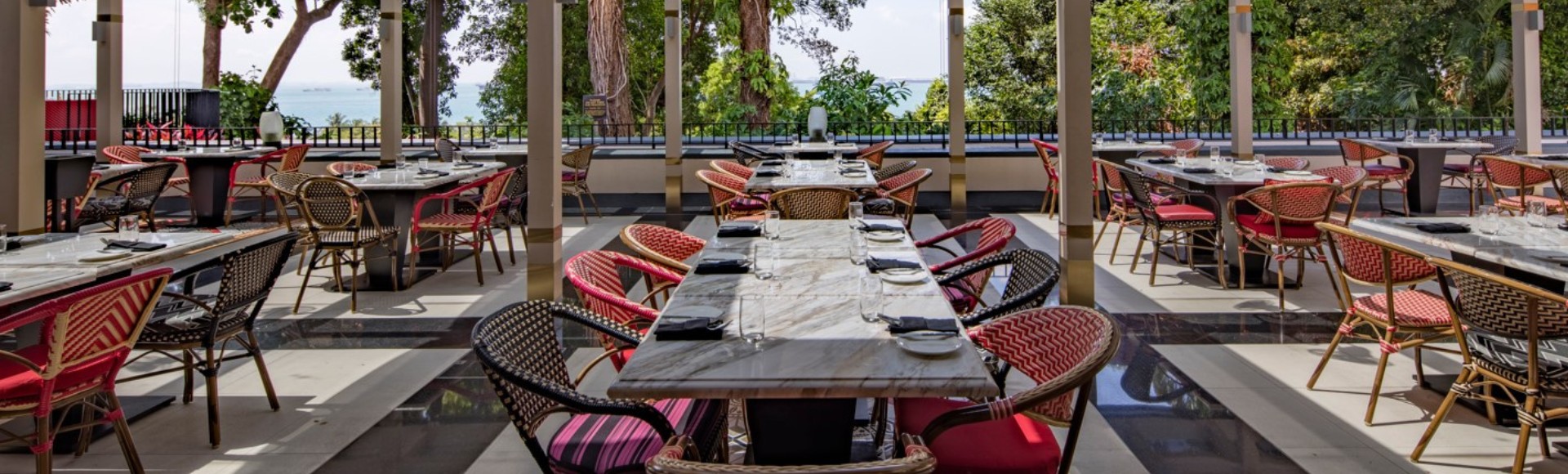 Outdoor restaurant with elegant marble-topped tables and red cushioned chairs, surrounded by lush greenery and a view of the ocean in the distance.