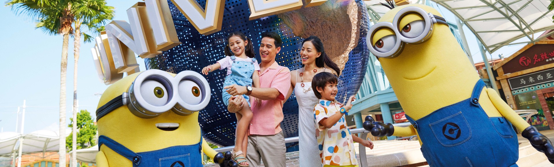 A joyful family poses with two large Minion characters at a theme park, near the 