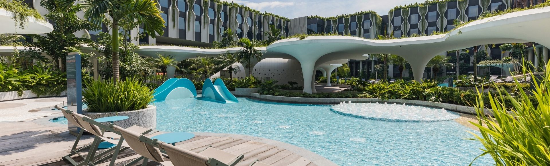 Pool area with modern architecture, featuring sun loungers, blue tables, lush greenery, and a playful slide, under a partly cloudy sky.