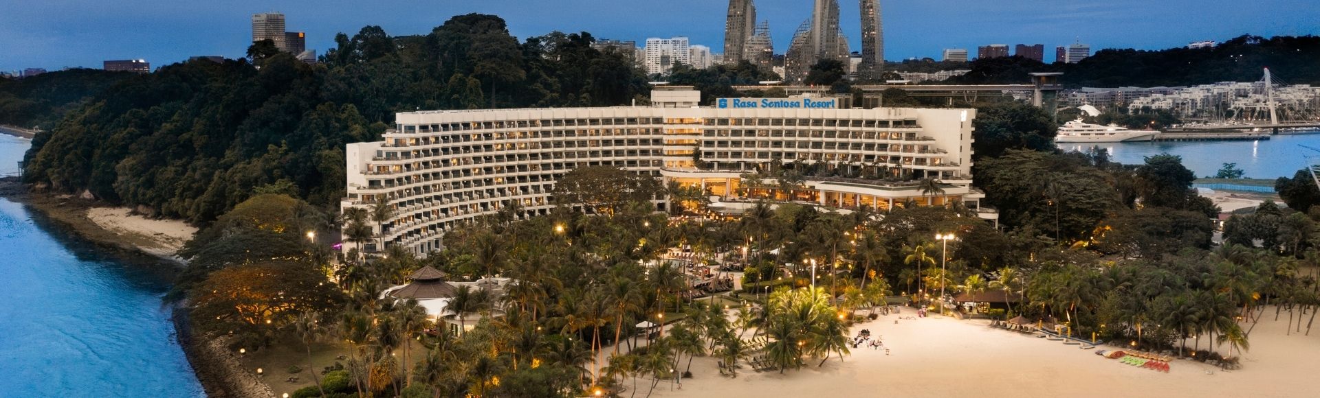 Aerial view of a beachfront resort with curved architecture, surrounded by lush greenery. The calm, blue ocean meets a sandy shore under a twilight sky.