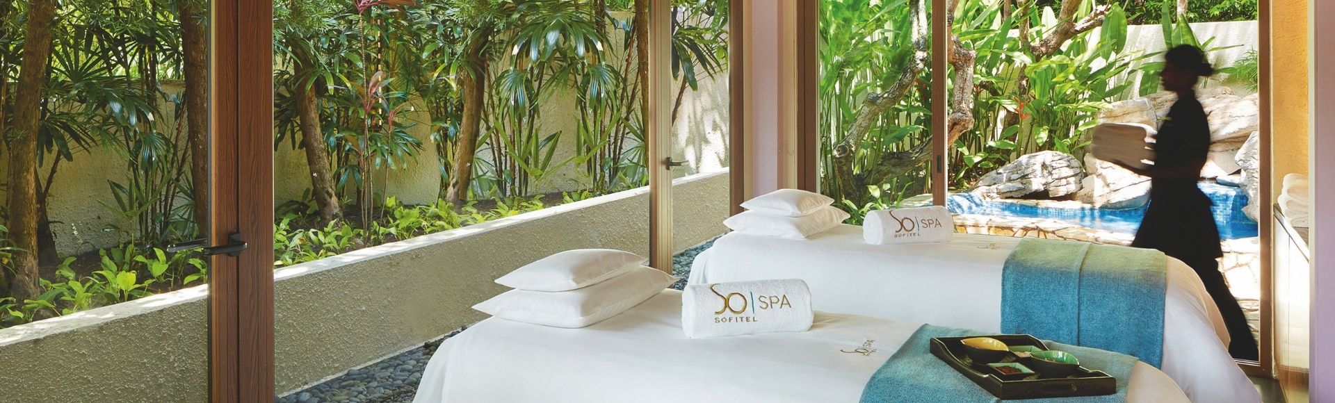 A serene spa room with two massage tables draped in white and blue linens, set beside large windows overlooking lush greenery. A staff member walks by.