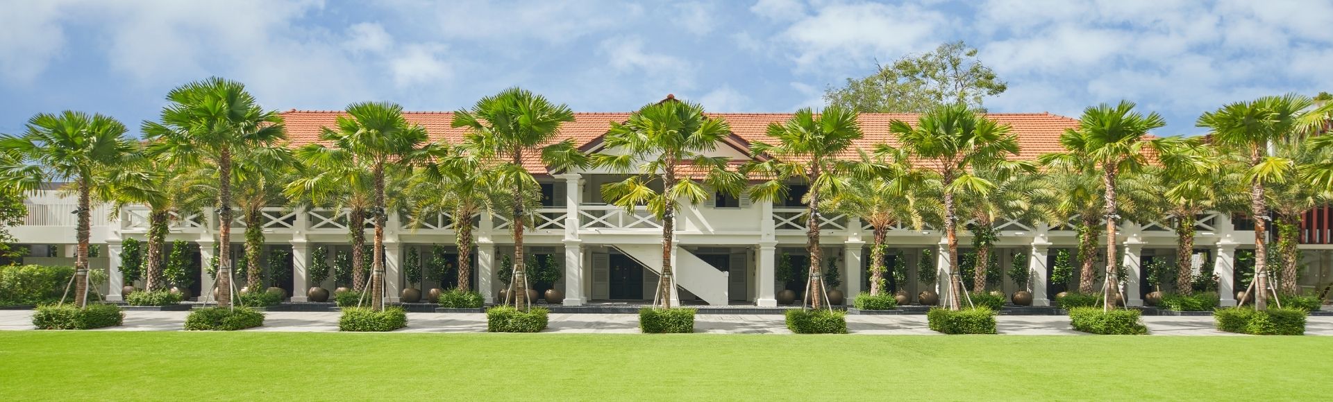 Two-story building with red-tiled roof, surrounded by palm trees. Lush green lawn in the foreground, clear blue sky above, conveying a serene tropical vibe.