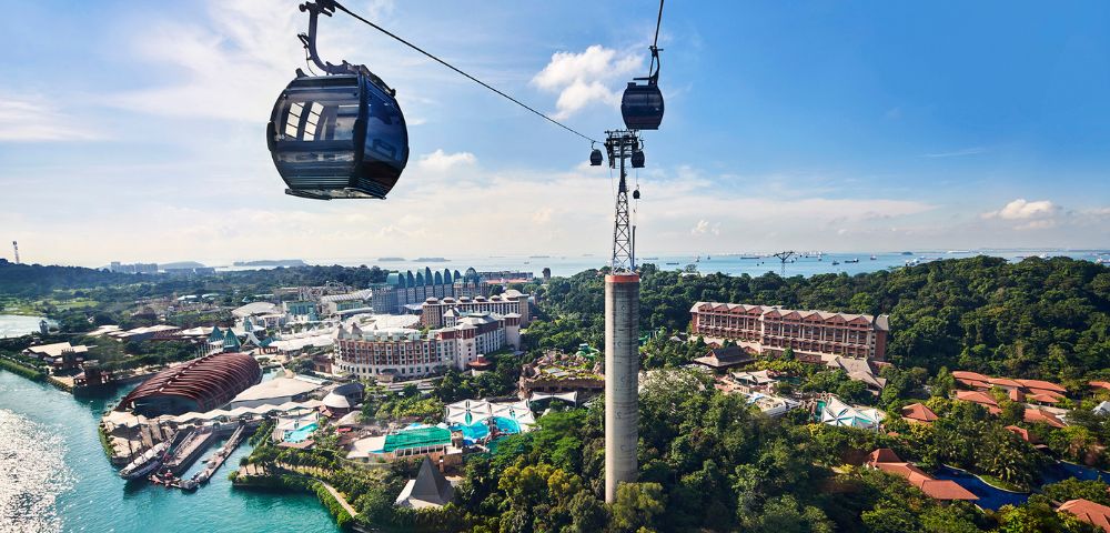 Two cable cars glide through the air over lush greenery and a vibrant resort; the sea and a blue sky with clouds complete the scenic view.
