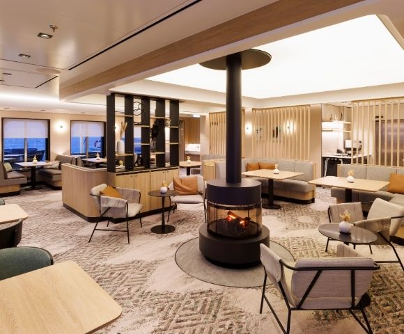 A modern lounge area with comfortable seating, tables, and a central fireplace.