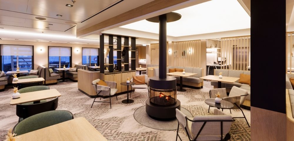 A modern lounge area with comfortable seating, tables, and a central fireplace.