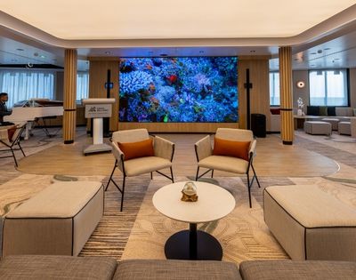 A spacious lounge area featuring a white grand piano with a musician playing, two modern armchairs with orange cushions, a round coffee table with a glass orb centerpiece, and a large LED screen displaying a vibrant coral reef.