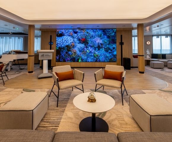 A spacious lounge area featuring a white grand piano with a musician playing, two modern armchairs with orange cushions, a round coffee table with a glass orb centerpiece, and a large LED screen displaying a vibrant coral reef.