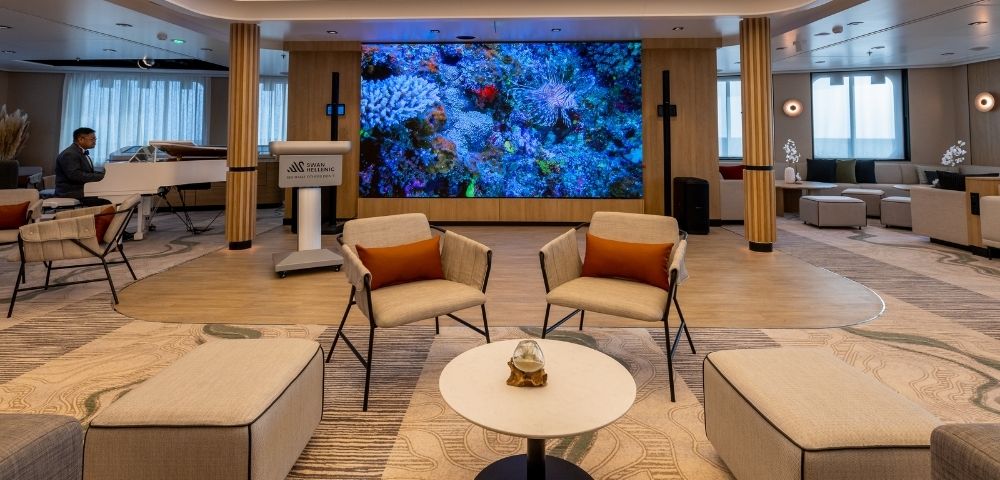 A spacious lounge area featuring a white grand piano with a musician playing, two modern armchairs with orange cushions, a round coffee table with a glass orb centerpiece, and a large LED screen displaying a vibrant coral reef.
