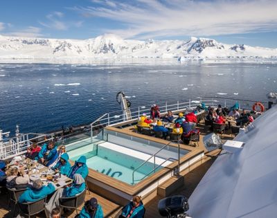 A cruise ship deck with people dining and relaxing near a small pool, with a backdrop of snow-capped mountains and icebergs in the ocean under a clear sky.