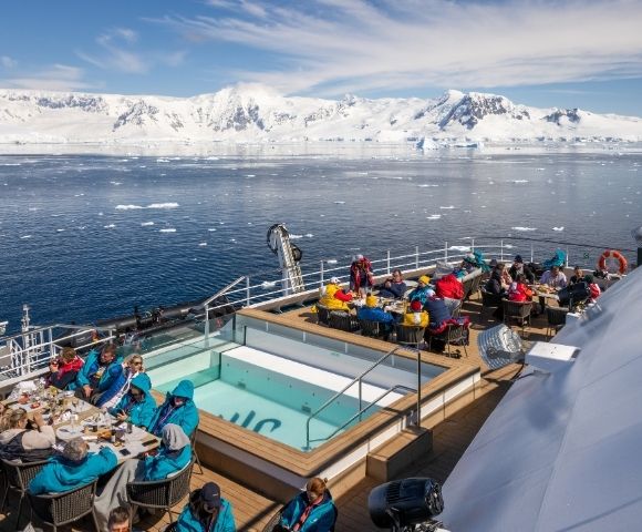 A cruise ship deck with people dining and relaxing near a small pool, with a backdrop of snow-capped mountains and icebergs in the ocean under a clear sky.