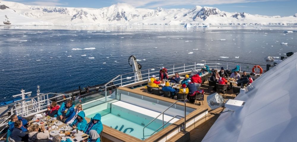 A cruise ship deck with people dining and relaxing near a small pool, with a backdrop of snow-capped mountains and icebergs in the ocean under a clear sky.