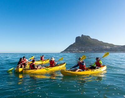 A group of people in bright yellow kayaks paddle on a calm blue sea under a clear sky, with a town nestled on a green mountain in the background.
