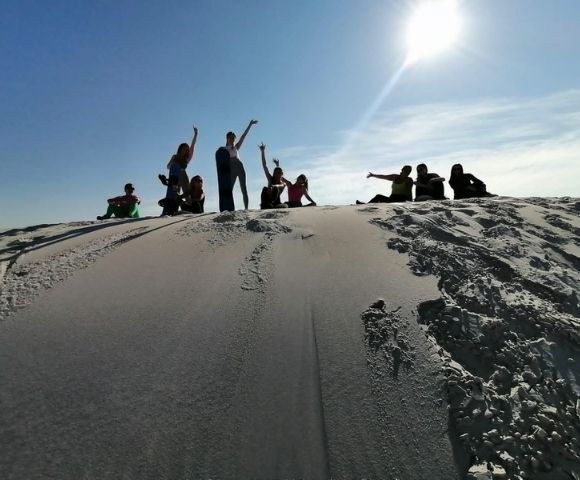 A group of people are posing and sitting on a sandy dune under a clear blue sky.