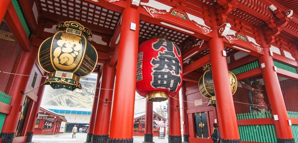 Large, ornate, traditional Japanese lanterns hang from the brightly painted red pillars of a temple entrance.