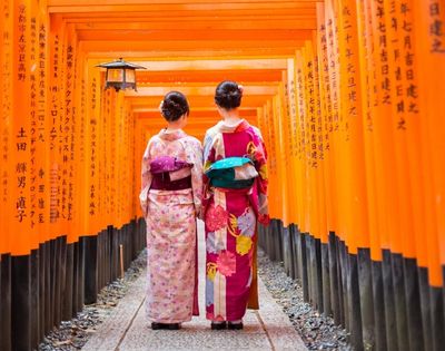 Two people in traditional Japanese kimonos walk through a tunnel of bright orange torii gates, with Japanese characters inscribed on the gates.