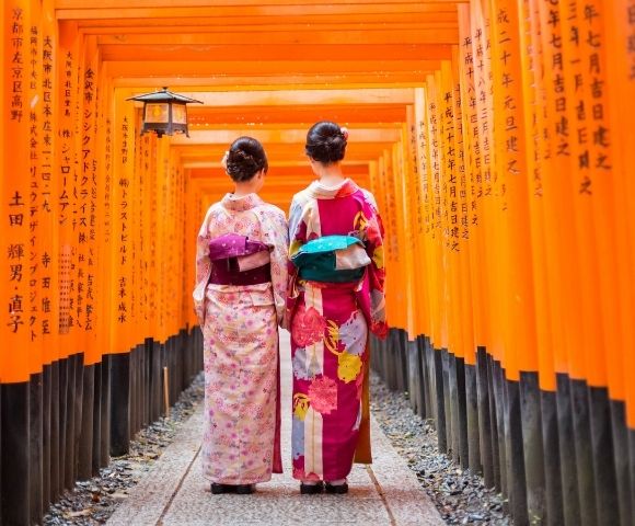 Two people in traditional Japanese kimonos walk through a tunnel of bright orange torii gates, with Japanese characters inscribed on the gates.