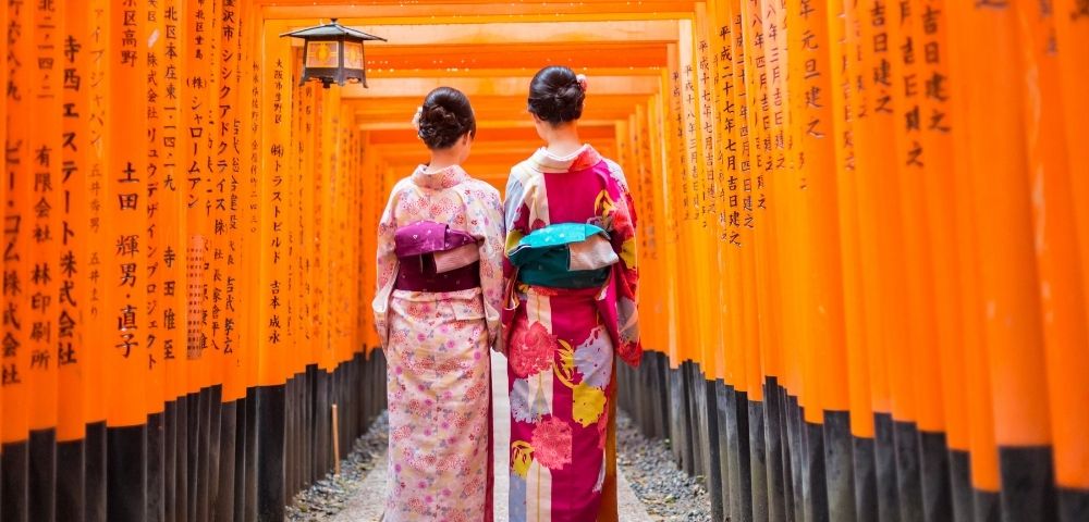 Two people in traditional Japanese kimonos walk through a tunnel of bright orange torii gates, with Japanese characters inscribed on the gates.