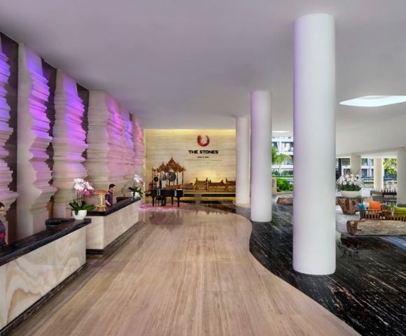 The lobby of a modern hotel with a unique textured wall illuminated by purple light, a grand piano, and prominent white columns.