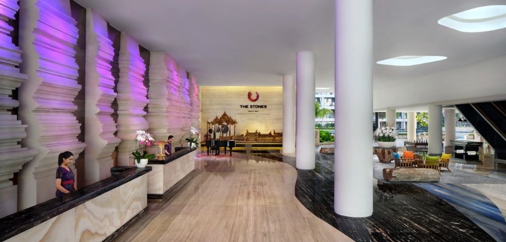 The lobby of a modern hotel with a unique textured wall illuminated by purple light, a grand piano, and prominent white columns.