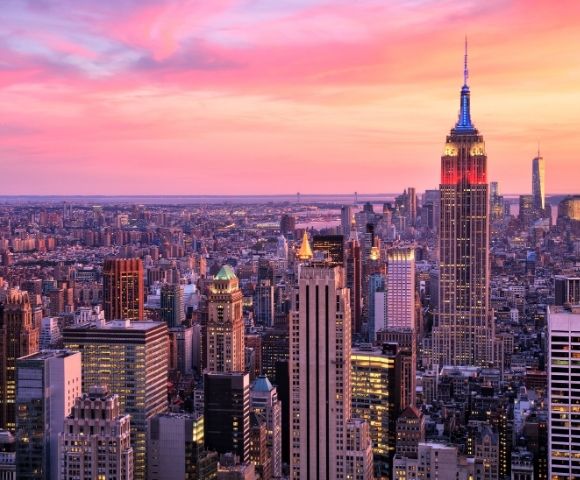 Panoramic aerial view of the New York City skyline at sunset, dominated by the illuminated Empire State Building showcasing red and blue lights, against a vibrant pink and orange sky.