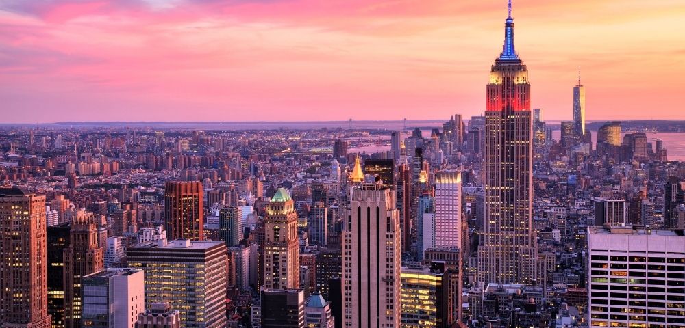 Panoramic aerial view of the New York City skyline at sunset, dominated by the illuminated Empire State Building showcasing red and blue lights, against a vibrant pink and orange sky.