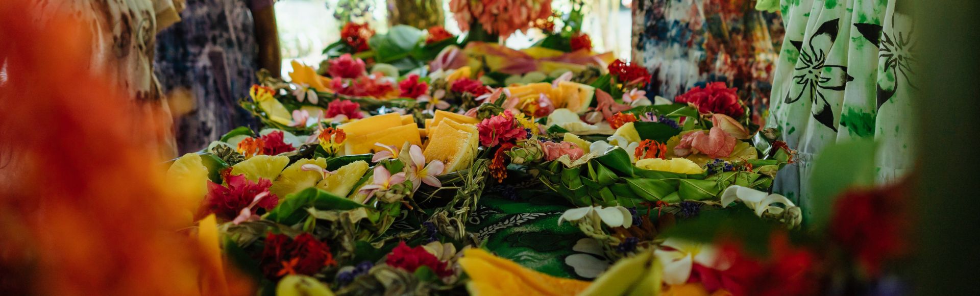 Tropical fruits like papaya and pineapple are artfully arranged among vibrant red and white flowers on lush greenery, conveying a festive and tropical vibe.