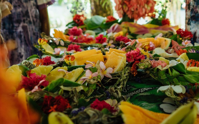Tropical fruits like papaya and pineapple are artfully arranged among vibrant red and white flowers on lush greenery, conveying a festive and tropical vibe.