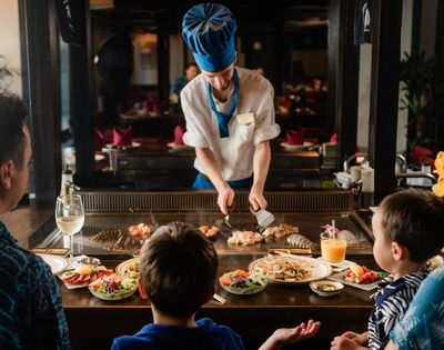 Chef in blue hat cooking seafood on teppanyaki grill for family dining at restaurant table with food and drinks.