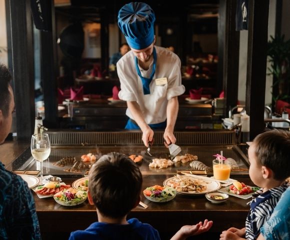 Chef in blue hat cooking seafood on teppanyaki grill for family dining at restaurant table with food and drinks.