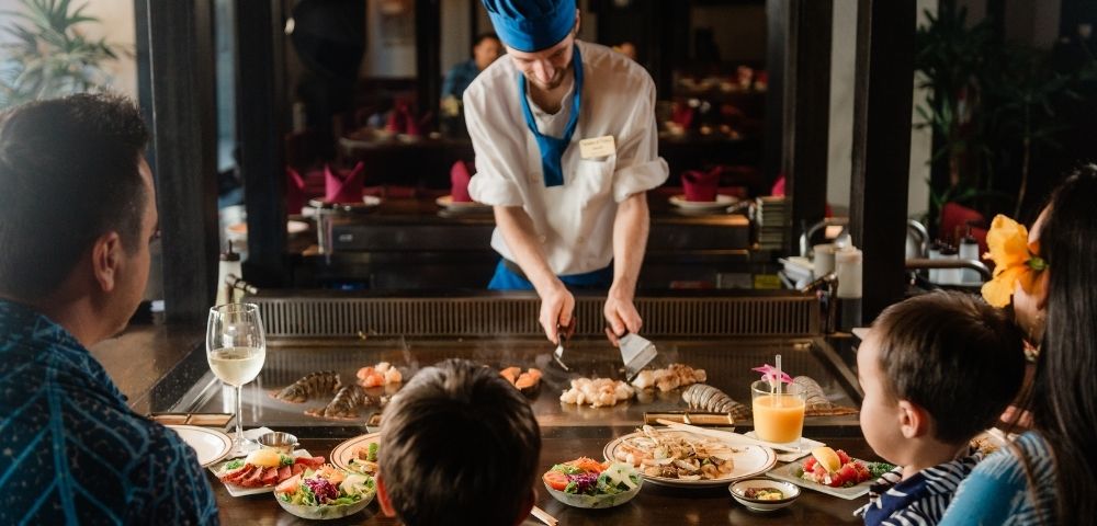 Chef in blue hat cooking seafood on teppanyaki grill for family dining at restaurant table with food and drinks.