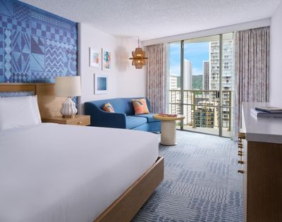 Modern hotel room with a large bed, blue accent wall, and a cozy seating area. Floor-to-ceiling windows show a cityscape view, creating a serene ambiance.