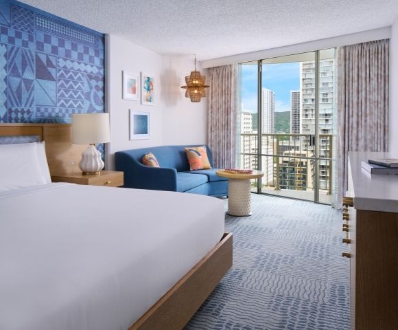Modern hotel room with a large bed, blue accent wall, and a cozy seating area. Floor-to-ceiling windows show a cityscape view, creating a serene ambiance.
