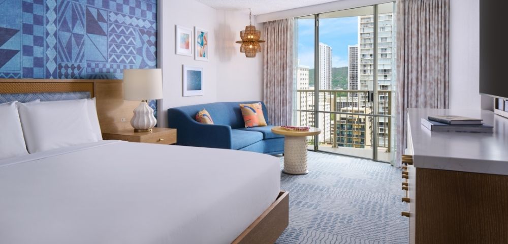 Modern hotel room with a large bed, blue accent wall, and a cozy seating area. Floor-to-ceiling windows show a cityscape view, creating a serene ambiance.