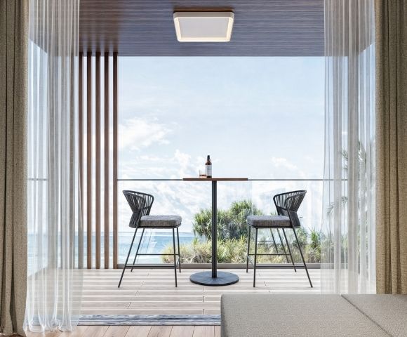 A contemporary balcony with two black wicker bar stools and a small circular table with a bottle and glass, overlooking the ocean.