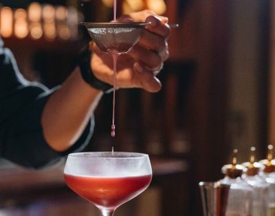 A hand carefully strains a deep red liquid into a glass, creating a sense of precision and focus. The background is softly blurred, highlighting a cozy atmosphere.