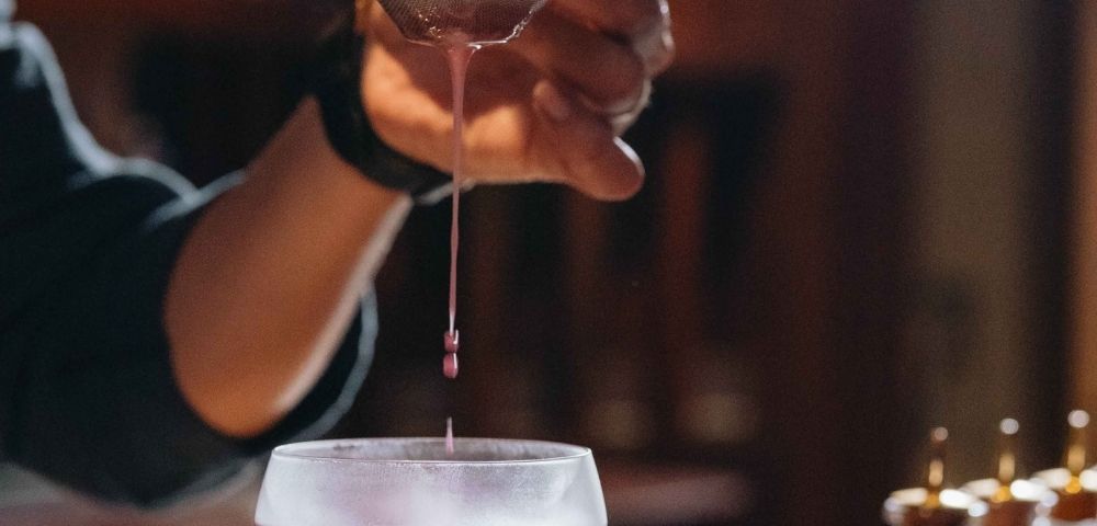 A hand carefully strains a deep red liquid into a glass, creating a sense of precision and focus. The background is softly blurred, highlighting a cozy atmosphere.