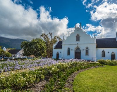 White Dutch Colonial style church with dark roof, gothic arched windows, and ornate gable, set against a backdrop of green trees and a mountain range under a dramatic cloudy blue sky, with a vibrant foreground of purple and white flowers bordering a lush green lawn.