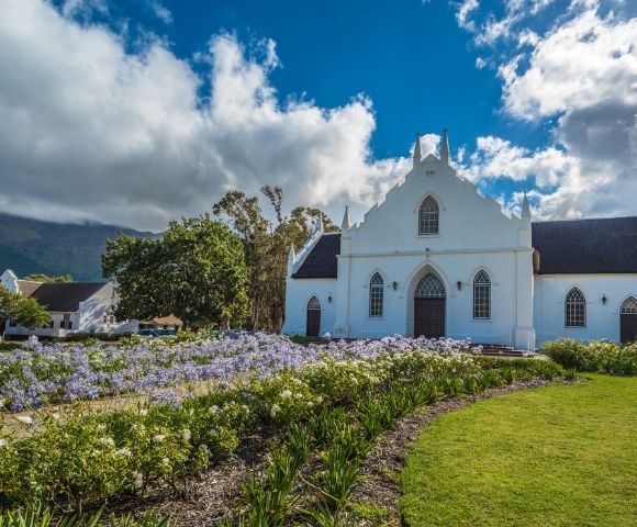 White Dutch Colonial style church with dark roof, gothic arched windows, and ornate gable, set against a backdrop of green trees and a mountain range under a dramatic cloudy blue sky, with a vibrant foreground of purple and white flowers bordering a lush green lawn.