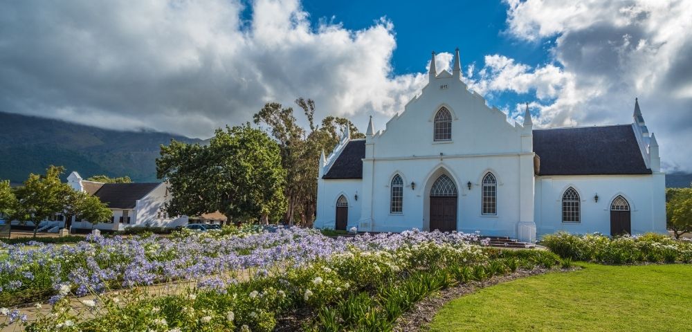 White Dutch Colonial style church with dark roof, gothic arched windows, and ornate gable, set against a backdrop of green trees and a mountain range under a dramatic cloudy blue sky, with a vibrant foreground of purple and white flowers bordering a lush green lawn.
