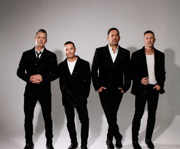 Four men stand side-by-side wearing matching sequined black blazers over white shirts and black pants.