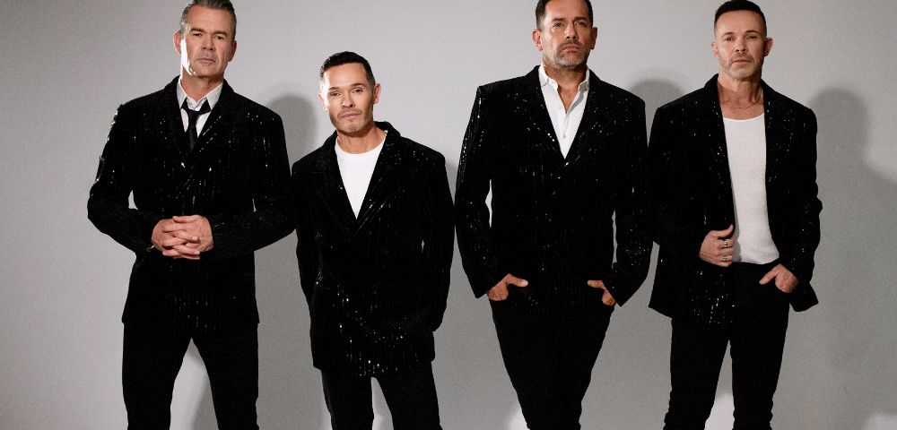 Four men stand side-by-side wearing matching sequined black blazers over white shirts and black pants.