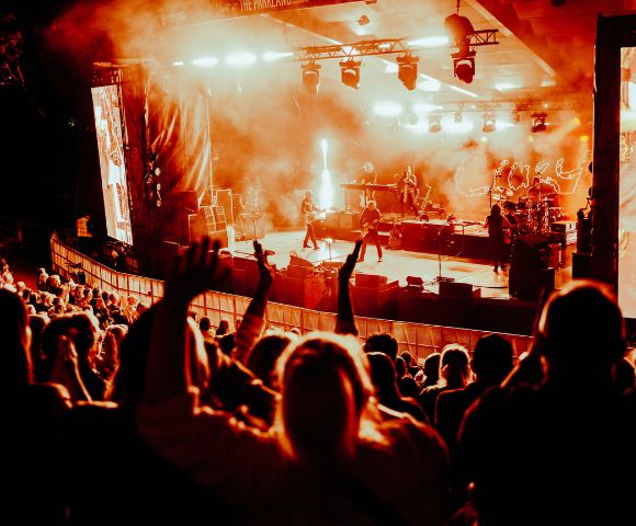 A band performs on a brightly lit stage with smoke effects, in front of a large, cheering crowd.