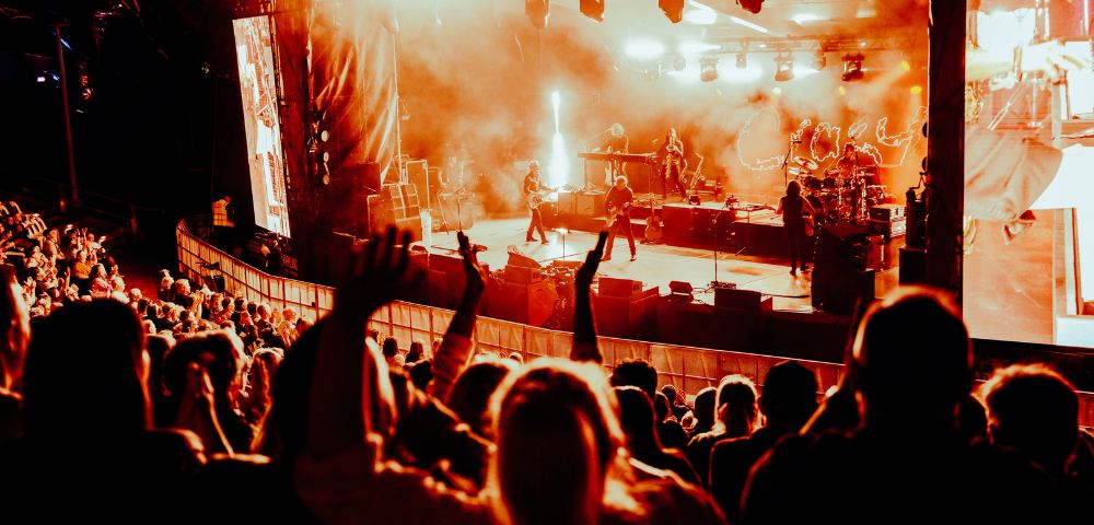 A band performs on a brightly lit stage with smoke effects, in front of a large, cheering crowd.