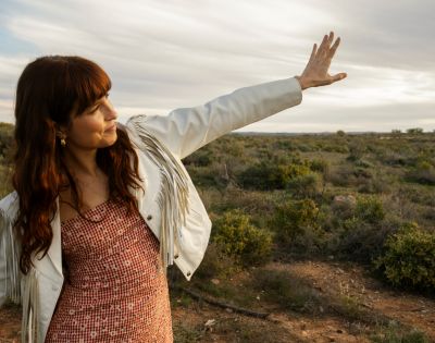 A woman with long brown hair wears a fringed white jacket and a red and white checkered dress as she holds her hand up to block the sun in a dry, brush-covered landscape under a cloudy sky.